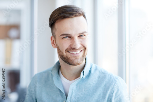 Head and shoulders of handsome young man looking at camera while posing by window in office or apartment, copy space