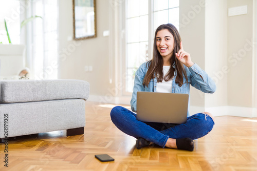Beautiful young woman sitting on the floor with crossed legs using laptop surprised with an idea or question pointing finger with happy face, number one