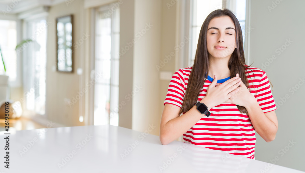 Beautiful young woman wearing casual stripes t-shirt smiling with hands on chest with closed eyes and grateful gesture on face. Health concept.