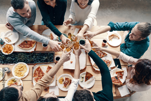 To our friendship! Top view of young people in casual wear toasting each other and smiling while having a dinner party indoors