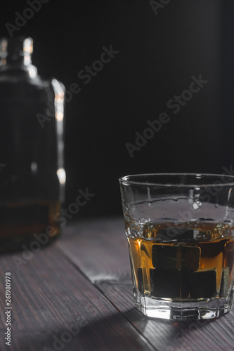 Vertical photo of a glass of whiskey and ice stones stands in the foreground on a wooden table in the background a fragment of a bottle