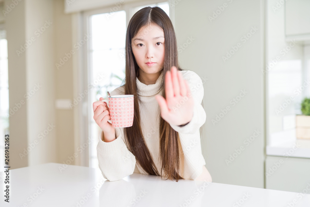 Beautiful Asian woman drinking a cup of coffee with open hand doing stop sign with serious and confident expression, defense gesture Beautiful Asian woman drinking a cup of coffee with open hand doing stop sign with serious and confident expression, defense gesture