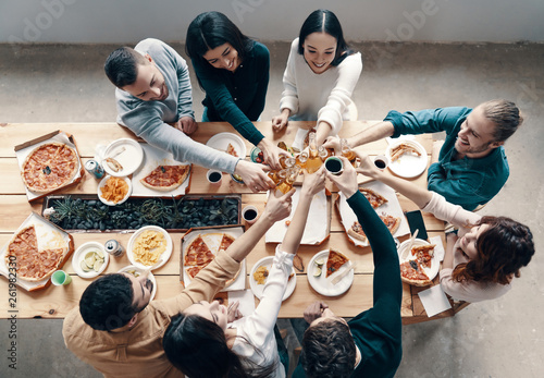 Cheers! Top view of young people in casual wear toasting each other and smiling while having a dinner party indoors