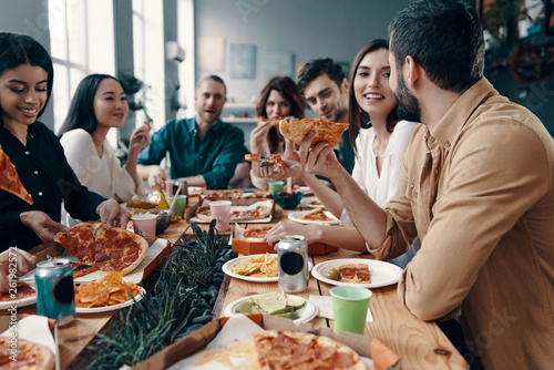 Holiday among friends. Group of young people in casual wear eating pizza and smiling while having a dinner party indoors