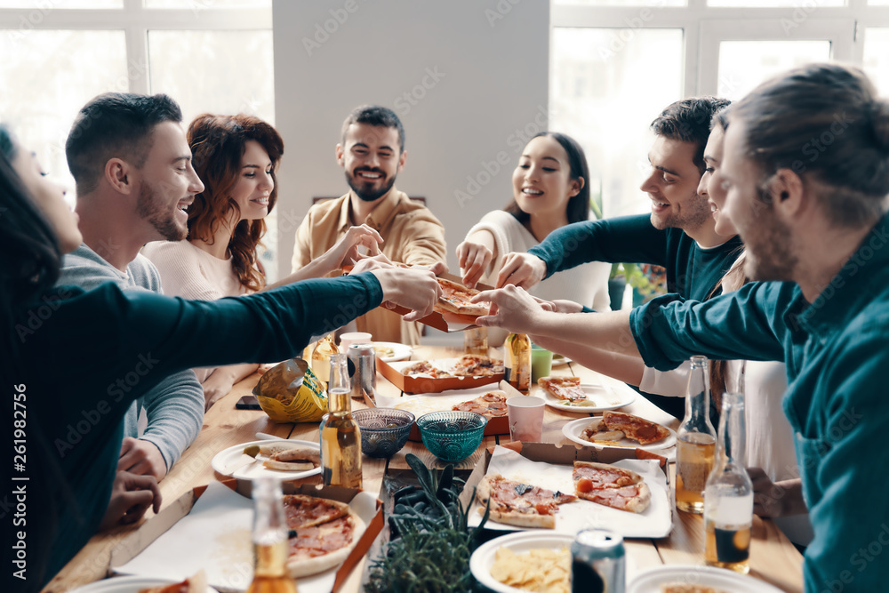 Hurry up to pick the best slice! Group of young people in casual wear picking pizza and smiling while having a dinner party indoors