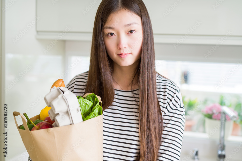 Beautiful Asian woman holding paper bag of fresh groceries with a confident expression on smart face thinking serious Beautiful Asian woman holding paper bag of fresh groceries with a confident expression on smart face thinking serious