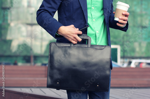 Cropped image businessman in casual wear tries to hold up cup of coffee to go, leather briefcase and smartphone at the same time against modern office building background photo