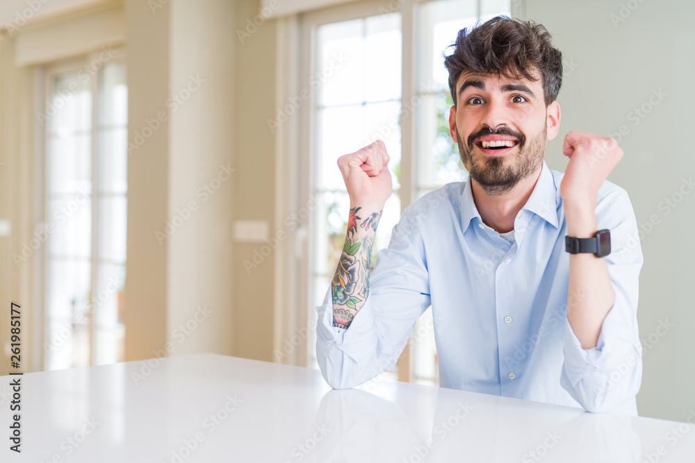 Young businesss man sitting on white table celebrating surprised and amazed for success with arms raised and open eyes. Winner concept. Young businesss man sitting on white table celebrating surprised and amazed for success with arms raised and open eyes. Winner concept.