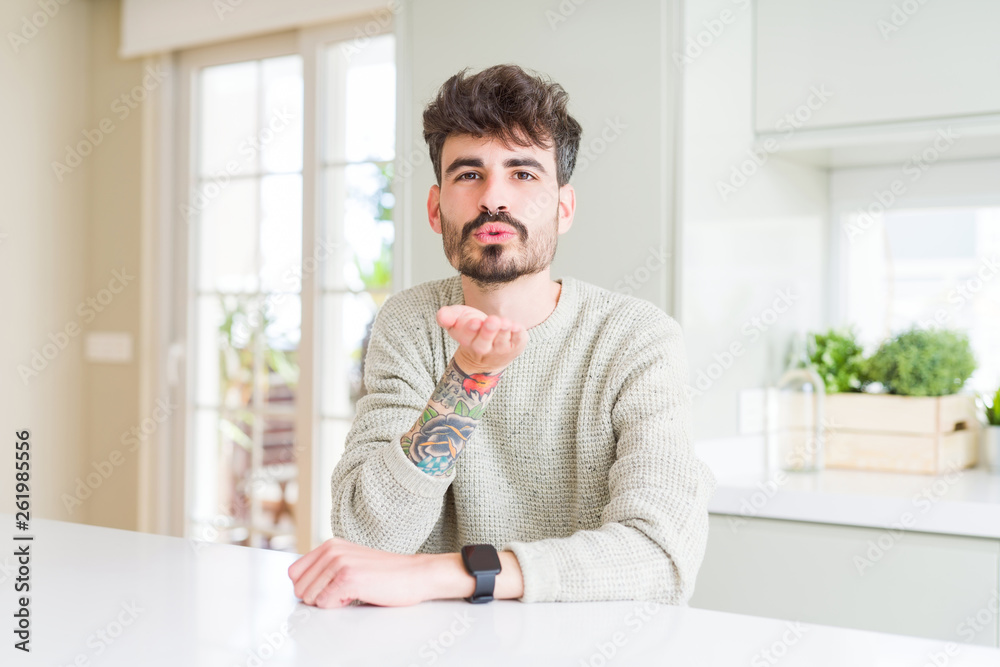 Young man wearing casual sweater sitting on white table looking at the camera blowing a kiss with hand on air being lovely and sexy. Love expression.