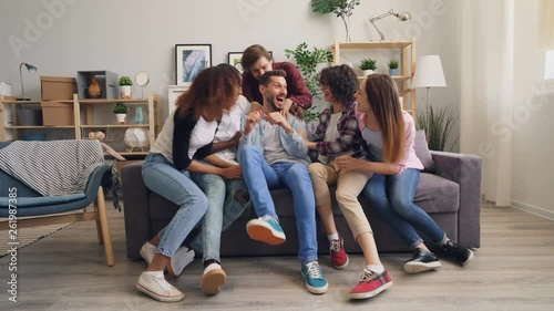 Sad handfsome guy is sitting on sofa alone then joyful friends are running to him hugging and laughing having fun and talking together. Friendship and house concept.
