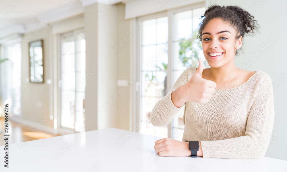Beautiful young african american woman with afro hair sitting on table at home doing happy thumbs up gesture with hand. Approving expression looking at the camera showing success. Beautiful young african american woman with afro hair sitting on table at home doing happy thumbs up gesture with hand. Approving expression looking at the camera showing success.