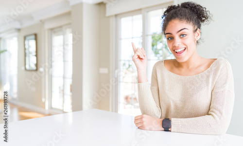 Beautiful young african american woman with afro hair sitting on table at home with a big smile on face, pointing with hand and finger to the side looking at the camera.