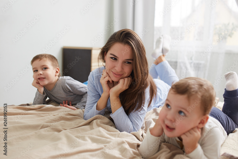Happy young mother dressed in light blue pajama lays with her two little sons holding their hands under their chins on the bed with beige blanket in the bedroom with big window Happy young mother dressed in light blue pajama lays with her two little sons holding their hands under their chins on the bed with beige blanket in the bedroom with big window