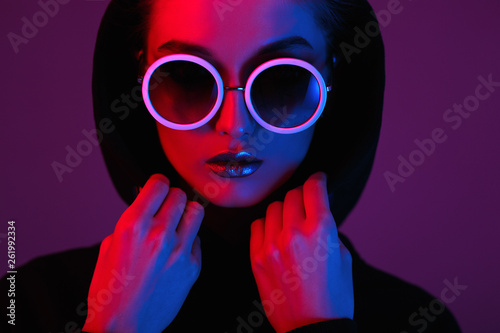 Portrait of fashion young girl in a black sweater with a hood and round sunglasses in red and blue neon light in the studio