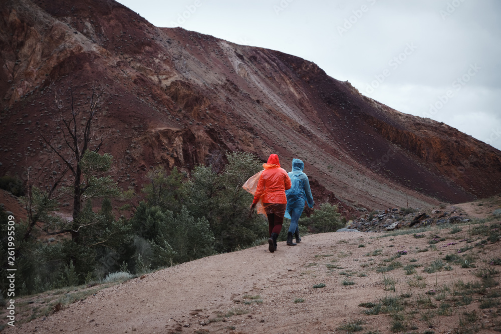 Two girls with excellent mood in raincoats travel. Rainy weather in the mountains prevents trekking. Go on a mountain trail.