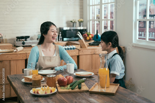 asian chinese young mother with children having breakfast in kitchen at home. happy parent kid relationship family. laughing woman housewife in apron with daughter wear uniform talking in morning.
