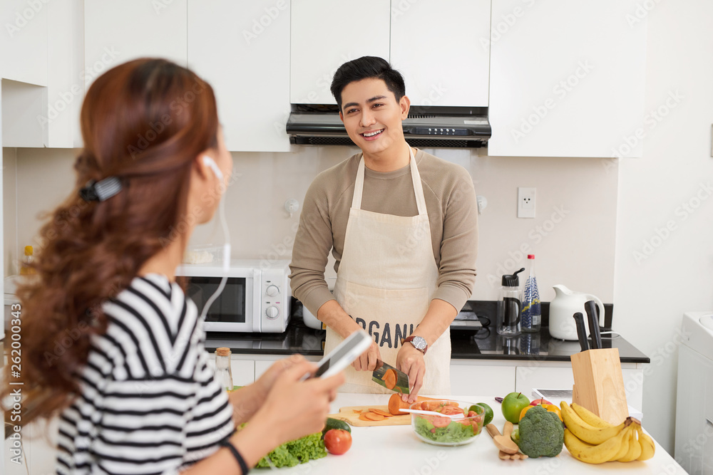 Beautiful young couple is cooking in kitchen at home.Man is cooking with woman is using a digital tablet and smiling. Beautiful young couple is cooking in kitchen at home.Man is cooking with woman is using a digital tablet and smiling.