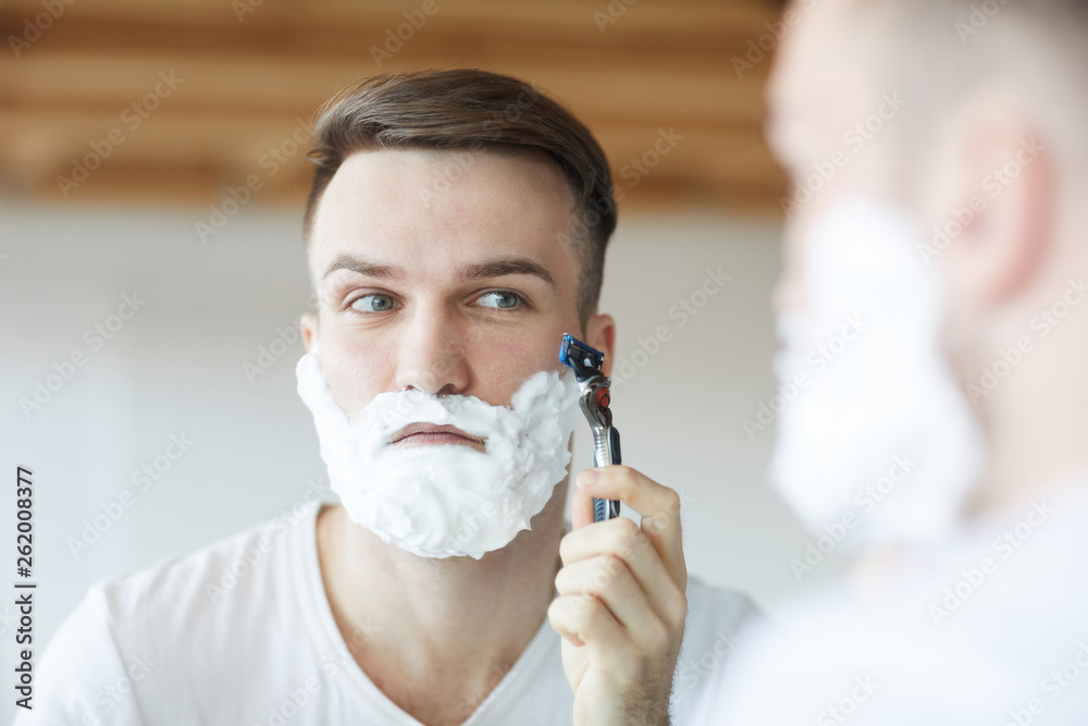 Head and shoulders portrait of handsome young man shaving in morning  looking at his reflection in mirror, copy space