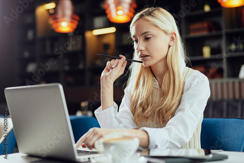 Young Caucasian blonde businesswoman dressed smart casual using laptop and holding pen while sitting in coffee shop. photo