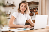 Image of elegant blond woman 20s wearing white t-shirt studying on laptop and writing down notes in apartment