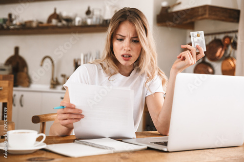 Image of serious blond woman holding plastic credit card while looking at paper documents in apartment