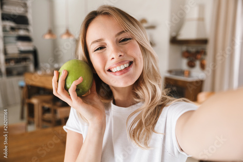 Image closeup of joyful blond woman laughing and holding green apple while taking selfie photo in living room
