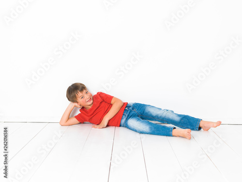 cute and blond boy with red shirt and blue jeans is posing on white wooden floor in front of white background