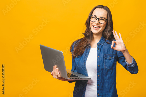 Portrait of a cheerful young girl holding laptop computer and showing ok isolated over yellow background. photo