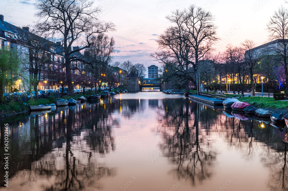 Amsterdam canal landscape at sunset with trees reflection in water and sky scraper in center and far perspective with boats parked along canal 