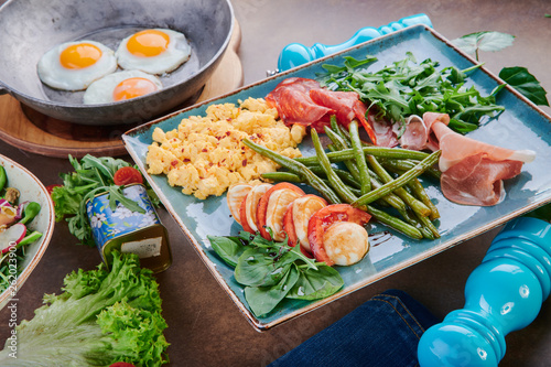 Scrambled eggs, slices of jamon and chorizo, asparagus bean and caprese on a beautiful blue plate and fried eggs on a beautiful brown table. Close up. Space