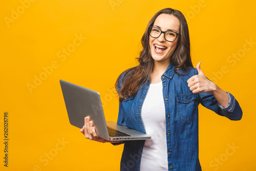 Portrait of a smiling young beautiful girl holding laptop computer and showing thumbs up isolated over yellow background.