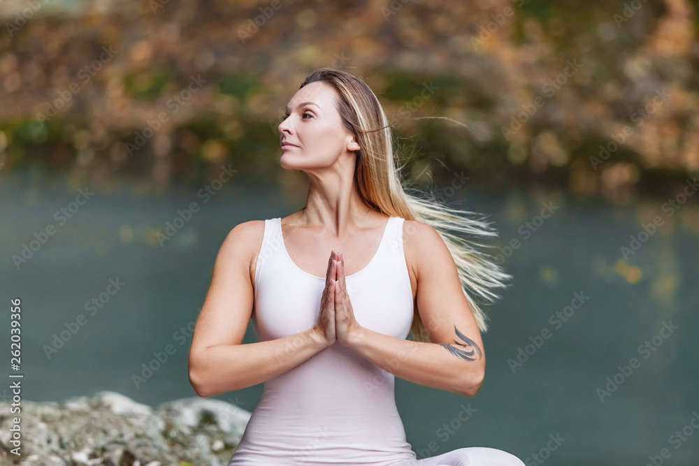 Pacified relaxed young beautiful girl blonde yoga lover sits on the big stone and meditate in the lotus position on the background of the picturesque green rocks and the waterfall stream
