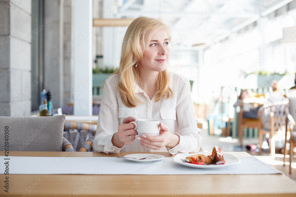 Cute young businesswoman sits at table and holds mug of tea next to dessert on the table and looks at passers-by walking along the city street.