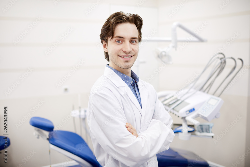 Waist up portrait of smiling young dentist posing in office next to dental chair, copy space Waist up portrait of smiling young dentist posing in office next to dental chair, copy space