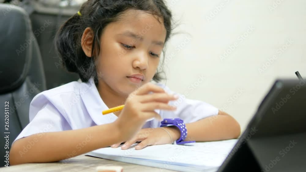Asian little girl in the Thai primary school uniform doing homework by using tablet smartphone for technology and education concept Asian little girl in the Thai primary school uniform doing homework by using tablet smartphone for technology and education concept