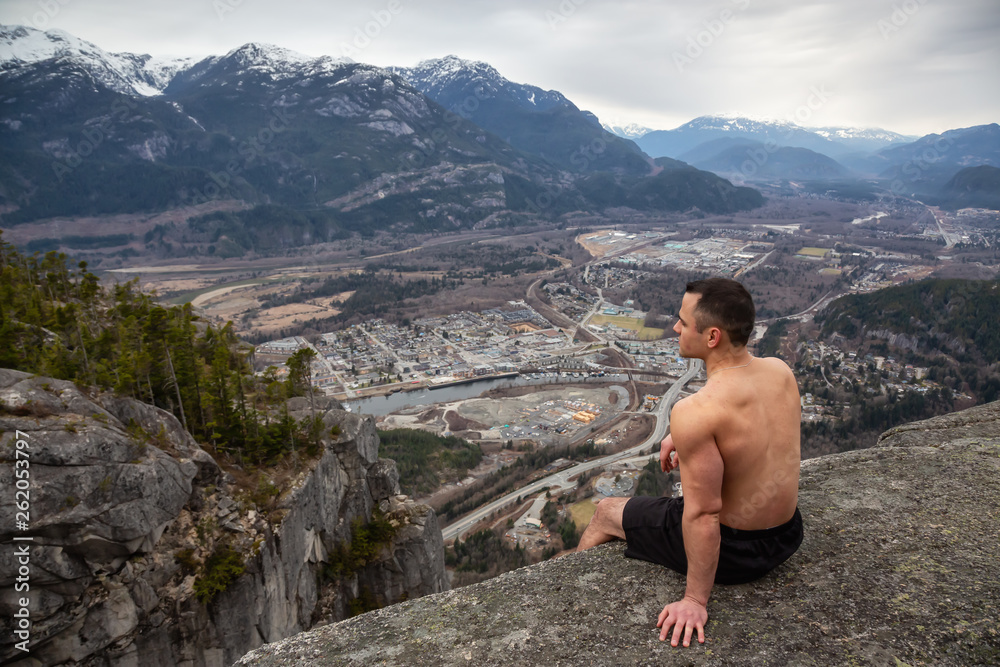 Fit and Muscular Caucasian Man is sitting at the cliff on top of a mountain and enjoying the scenery. Taken in Squamish, North of Vancouver, BC, Canada. Fit and Muscular Caucasian Man is sitting at the cliff on top of a mountain and enjoying the scenery. Taken in Squamish, North of Vancouver, BC, Canada.