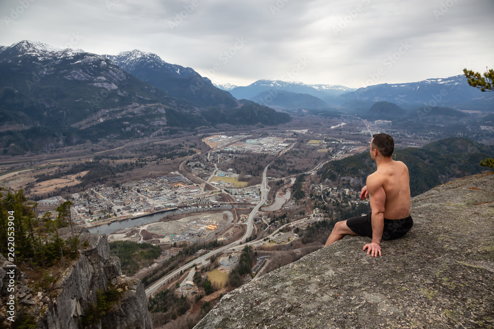 Fit and Muscular Caucasian Man is sitting at the cliff on top of a mountain and enjoying the scenery. Taken in Squamish, North of Vancouver, BC, Canada.