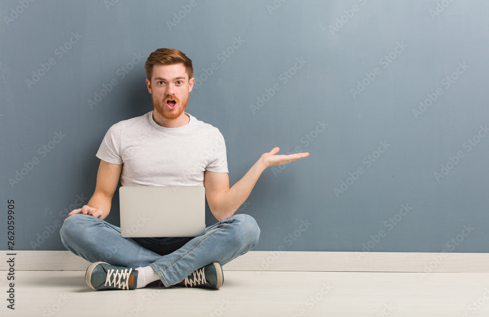 Young redhead student man sitting on the floor holding something on palm hand. He is holding a laptop. Young redhead student man sitting on the floor holding something on palm hand. He is holding a laptop.