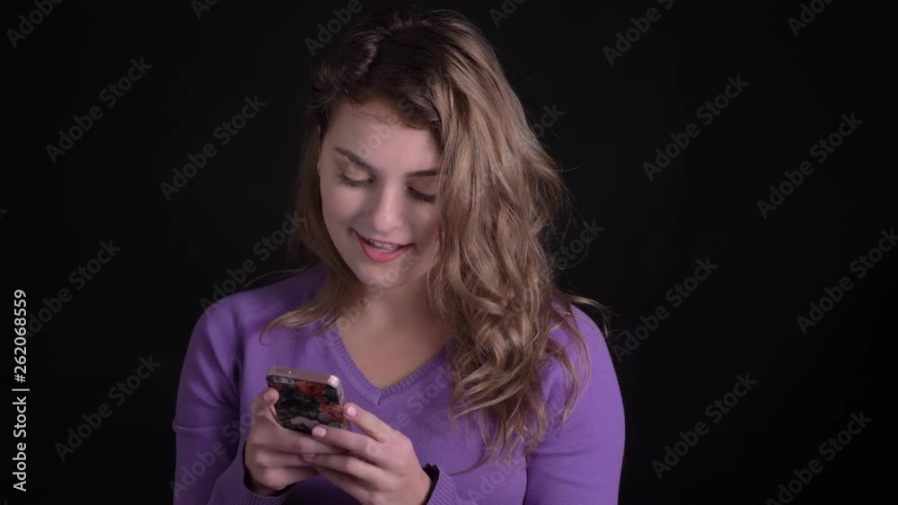 Portrait of glad woman watching with kind smile into smartphone on black background.