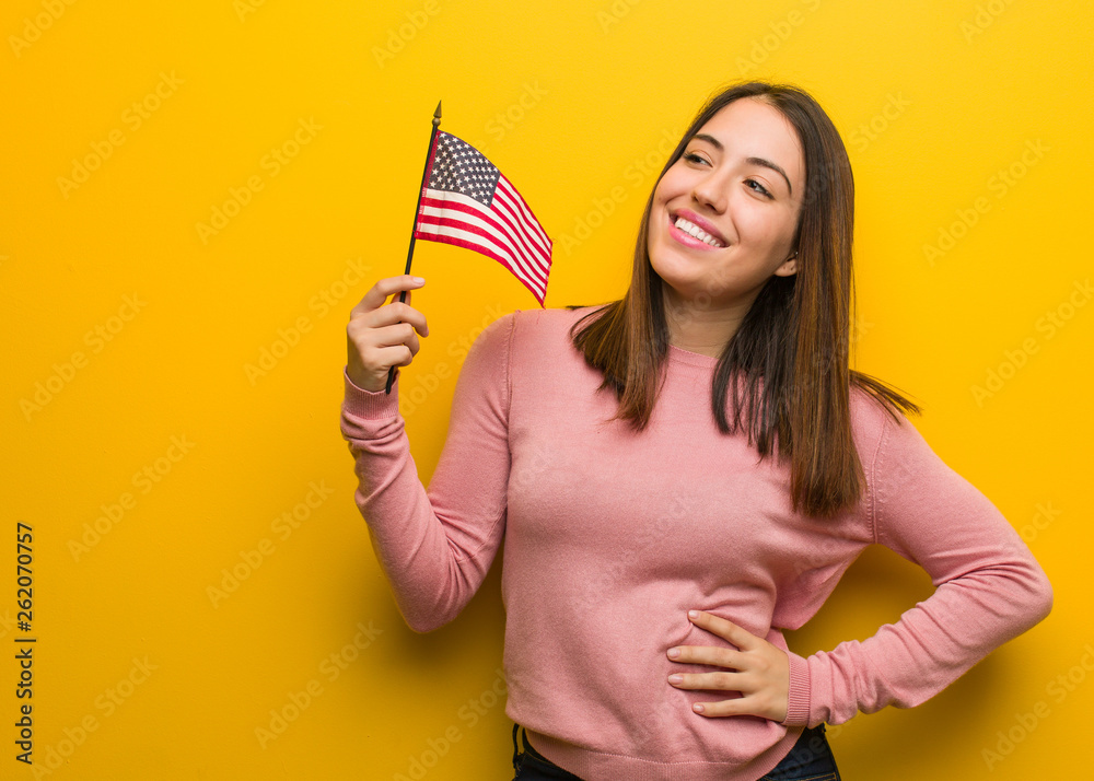 Young cute woman holding an united states flag smiling confident and crossing arms, looking up