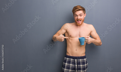 Young shirtless redhead man surprised, feels successful and prosperous. He is holding a coffee mug.