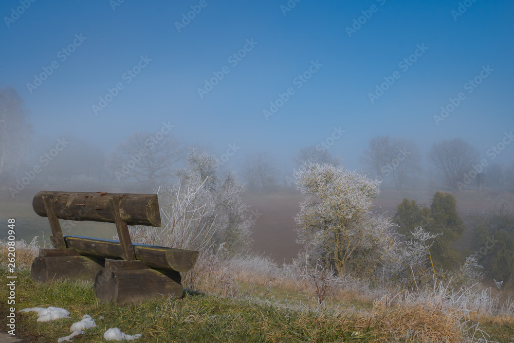 Early morning view to the old wooden bench on foreground and a field filled with fog with grass and bushes in frost on the background