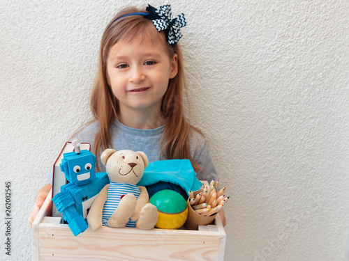 Cute little girl holds wooden box with donations: children's toys, stationery and children's clothing. Concept: you can help, they need help. Background, free space for inscription. 