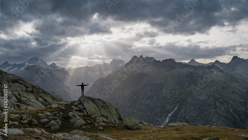 A boy on a trekking trip enjoys an incredible view of sun rays breaking through thick dark clouds over a valley, posing in "Jesus" or christian cross pose with half raised arms