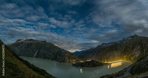 Night panorama with long exposure of blurred moving clouds and still stars on night sky taken in Swiss Alps on Grimsel lake.
