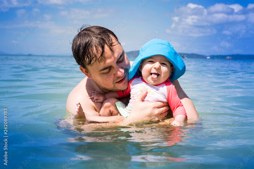 Child shows tongue, she is enjoy. Happy laughing infant baby with dad having fun in the sea. Father teaches daughter to swim and water.