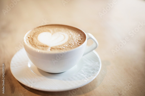White cappuccino cup with with latte art heart on light brown wood background lit by bright morning sunlight