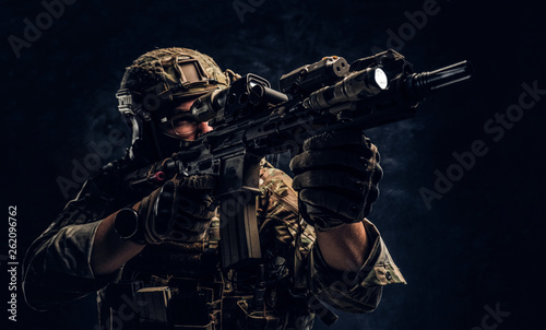 Close-up studio photo against a dark wall. The elite unit  special forces soldier in camouflage uniform holding an assault rifle with a laser sight and aims at the targe