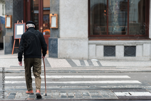An elderly male with a walking stick slowly walks across a crosswalk in the middle of a city alone