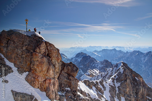 Golden summit cross and tiny climbers on top of Zugspitze peak with snowy climbing route Jubilaumsgrat ridge in background, Wetterstein range Bavarian / Northern Limestone Alps Bayern Germany Europe